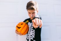 Menino sério em traje de esqueleto preto segurando Halloween Jack O Lanterna abóbora e apontando para a câmera enquanto está de pé contra o fundo branco — Fotografia de Stock