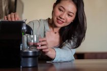 Junge Frau gegen Kaffeemaschine, die in Hausküche Heißgetränk mit Schaum ins Glas gießt — Stockfoto