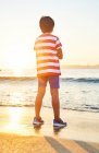 Вид ззаду на повну довжину невпізнаваного хлопчика, що стоїть на мокрому піщаному березі, омитому махаючи блакитним морем на заході сонця — стокове фото