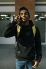 Красавчик афроамериканец хипстер в шляпе и толстовке улыбается и смотрит в камеру, разговаривая по смартфону на городской улице — стоковое фото
