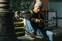 Содержание афроамериканский хипстер мужчина с рюкзаком сидит на каменной границе на улице и сообщения на мобильный телефон — стоковое фото