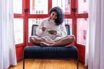 Full body of barefoot latin female sitting with crossed legs on chair and eating soup from bowl — Stock Photo