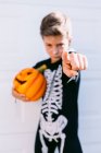 Niño serio disfrazado de esqueleto negro sosteniendo Halloween Jack O Linterna calabaza y apuntando a la cámara mientras está de pie contra el fondo blanco - foto de stock