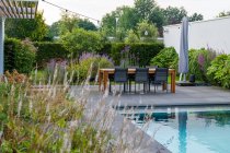 Tavolo da pranzo con sedia e lettini in legno posizionati vicino alla piscina nel cortile della costosa villa contemporanea in stile minimalista nella giornata di sole — Foto stock