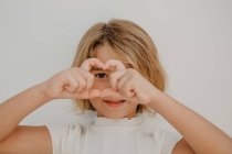 Criança amigável com cabelo castanho demonstrando gesto de amor com as mãos enquanto cobre o olho e olhando para a câmera — Fotografia de Stock