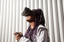 Jeune femme afro-américaine excitée dans un casque VR en utilisant un contrôleur tout en divertissant et en jouant à un jeu virtuel contre un mur rayé gris — Photo de stock