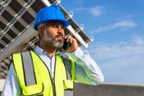 From below middle aged Hispanic foreman in in hardhat and waistcoat speaking on smartphone looking away while standing near solar power station — Stock Photo