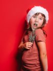 Bonito menina surpreendida em roupas casuais e chapéu de Papai Noel segurando galho de abeto e olhando para a câmera contra o fundo vermelho — Fotografia de Stock
