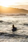 Rückansicht einer unkenntlichen jungen Frau mit Surfbrett, die während des Sonnenuntergangs am Strand in Asturien ins Meer steigt — Stockfoto