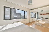 Moderne Schlafzimmereinrichtung mit Bett gegen Spiegelschrank und Fenster im Haus an sonnigen Tagen — Stockfoto