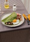 Homemade breakfast of spinach pancakes with bacon, egg and mushrooms served on a white plate with a salt and oil shaker on a checkered tablecloth. — Stock Photo