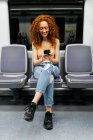 Donna interessata con i capelli ricci in jeans strappati messaggistica di testo sul cellulare durante il viaggio in treno di giorno — Foto stock