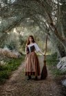 Bruxa séria de vestido de pé com livro mágico de feitiços e vassoura na estrada na floresta e olhando para a câmera — Fotografia de Stock