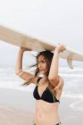 Smiling young female athlete in swimwear with flying hair carrying surfboard on head looking forward on ocean coast — Stock Photo
