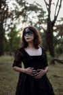 Мистическая ведьма в длинном черном платье с раскрашенным лицом, смотрящая в темные мрачные леса — стоковое фото