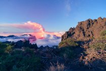 Nächtliche Landschaft mit einem ausbrechenden Vulkan im Hintergrund und einem Wolkenmeer, das die Berge in einer sternenklaren Nacht von einem bewachsenen und felsigen Berg bedeckt. Cumbre Vieja Vulkanausbruch auf La Palma Kanarische Inseln, Spanien 2021 — Stockfoto