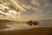 Scenic landscape of wet sandy shore with rocky formations under cloudy sky with sunlight in evening on beach Playon de Bayas in Asturais Spain — Stock Photo