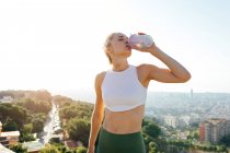 Atleta donna magra con mano sull'anca e occhi chiusi che beve acqua dalla bottiglia durante la pausa dall'allenamento in città — Foto stock