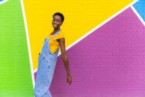 Vista lateral de la hembra afroamericana feliz sonriendo mientras salta por encima del suelo cerca de la pared brillante - foto de stock