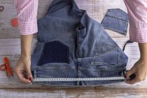 Alto ângulo de cultura costureira feminina anônima medindo cintura de jeans em mesa de madeira durante o dia — Fotografia de Stock