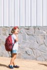 Side view of schoolchild with backpack touching lips on pavement while looking forward in sunlight — Stock Photo