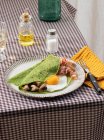 Petit déjeuner maison de crêpes d'épinards avec bacon, oeuf et champignons servi sur une assiette blanche avec une salière et une huileuse sur une nappe à carreaux. — Photo de stock