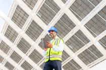 Niedriger Winkel eines glücklichen hispanischen Ingenieurs mittleren Alters in Uniform mit Tablet, der unter einem Solarkraftwerk wegschaut — Stockfoto