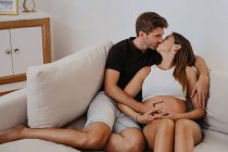 Чоловік цілується і приймає живіт очікуваної коханої жінки під час відпочинку на дивані у вітальні — стокове фото