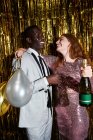 Дружелюбная молодая женщина с бутылкой шампанского и воздушными шарами обнимает любимого афроамериканца во время вечеринки — стоковое фото