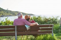 Вид ззаду на невпізнавану старшу пару, яка сидить, дивлячись один на одного на дерев'яній лавці і насолоджуючись літнім днем на березі ставка — стокове фото