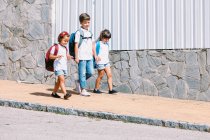 Школьник с рюкзаком разговаривает с подругами во время прогулки по мощеной тротуару у каменной стены при солнечном свете — стоковое фото