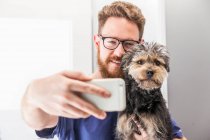 Médico veterinário masculino positivo tomando auto retrato com Yorkshire Terrier lambendo bochecha na clínica veterinária — Fotografia de Stock