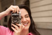 Молодая счастливая женщина с длинными каштановыми волосами, фотографирующая на старомодную фотокамеру на улице города — стоковое фото
