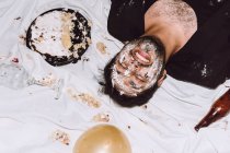 Drunk laughing male in smashed birthday cake lying near empty bottles from beer and balloons with eyes closed — Stock Photo