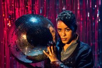 Trendy young ethnic female looking at camera while holding mirror ball in nightclub — Stock Photo