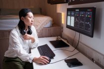 Вид сбоку концентрированной азиатской женщины, работающей на компьютере с графиками, показывающими динамику изменения стоимости криптовалют на удобном рабочем месте — стоковое фото