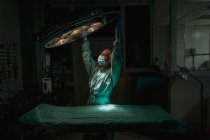Veterinário feminino jovem em uniforme cirúrgico e máscara estéril olhando para cima ao ajustar a lâmpada acima têxtil médica na clínica — Fotografia de Stock