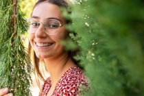 Весела молода жінка з коричневим волоссям в окулярах стоїть серед зелених гілок і дивиться на камеру в денне світло — стокове фото