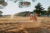 Side view of happy toddler child with toy sitting looking at camera in plastic bath while playing with water in countryside — Stock Photo