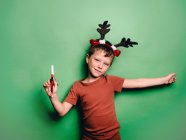 Boy wearing reindeer horns headband and festive party blower standing against green background and looking at camera — Stock Photo