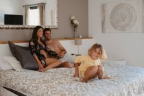 Fröhliches Kind im Schlafanzug hat Spaß im Bett gegen schwangere Mutter, die mit Ehemann im Haus interagiert — Stockfoto