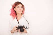Photographe féminine pensive avec des cheveux roses tenant un appareil photo professionnel dans ses mains dans la salle de lumière — Photo de stock