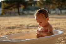 Niño pequeño con juguete sentado en un baño de plástico mientras juega con agua en el campo - foto de stock