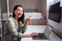 Visão lateral do conteúdo jovem blogueira étnica na mesa com netbook e café olhando para a câmera no quarto da casa — Fotografia de Stock