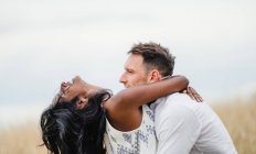 Vista lateral do homem sorridente abraçando namorada indiana em pé no campo sob céu nublado — Fotografia de Stock