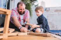 Barbudo padre enseñando hijo con martillo trabajando con madera mientras está sentado en el paseo marítimo en fin de semana - foto de stock