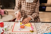 Crop unrecognizable kid in checkered dress with toy knife cutting fried eggs on chopping board while playing in house — Stock Photo