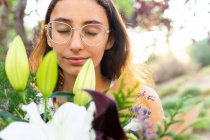 Crop mindful jovem fêmea com olhos fechados em óculos desfrutando aroma de buquê floral florescente na cidade no fundo borrado — Fotografia de Stock