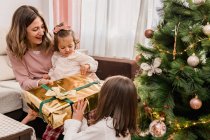 Mère joyeuse avec fille enfant en bas âge passant boîte cadeau à fille contre sapin décoré pendant les vacances du Nouvel An dans la maison — Photo de stock