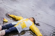 Alto ángulo de contenido Niño asiático en botas de goma y slicker acostado en charco ondulado en día lluvioso - foto de stock